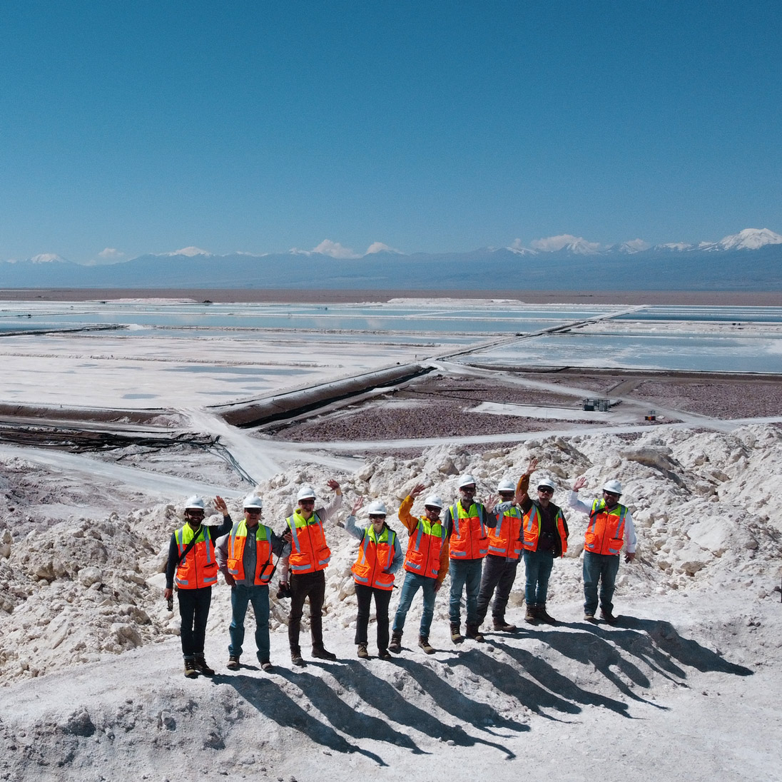 Eine Gruppe von Personen in Schutzkleidung steht vor einem weitläufigen Salzsee mit Verdunstungsbecken in bergiger Umgebung.