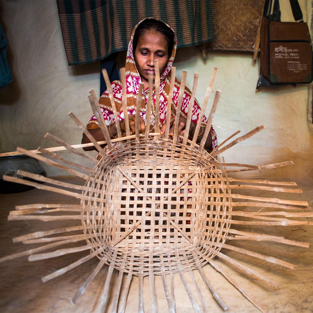 Eine Frau sitzt in einem Raum und flechtet mit den Händen einen großen Korb aus Holzstreifen.